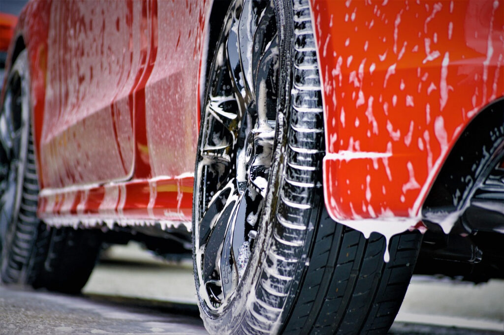 Close-up of a red car being washed during exterior detailing with soap foam covering the paint and wheels
